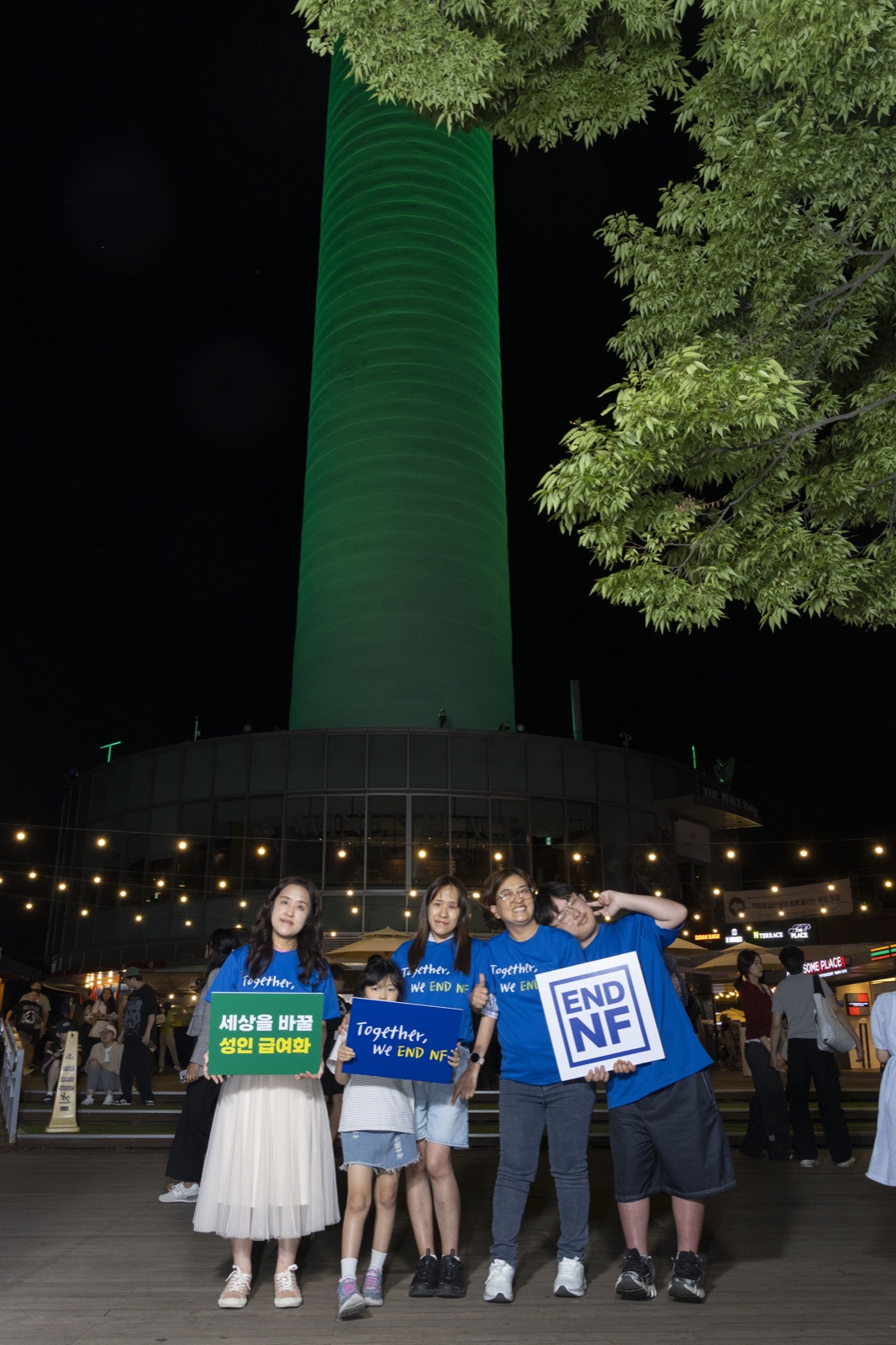 남산서울타워가 파란빛으로 점등된 야경. 환우 두 명이 'Together, We END NF' 메시지 보드를 들고 우러러보고 있다.