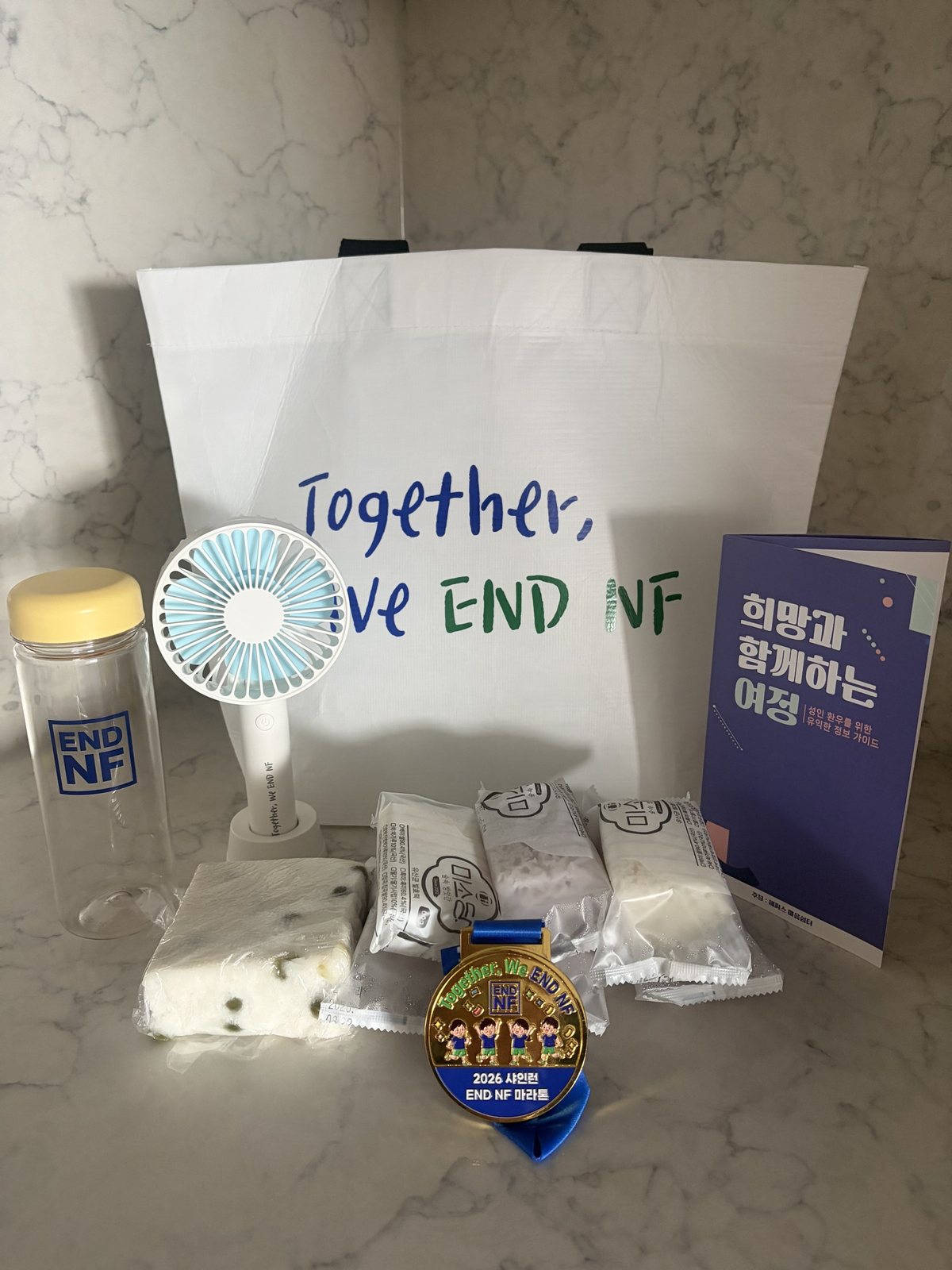 Together, We END NF 에코백과 END NF 물통, 손선풍기, 떡, 2026 샤인런 END NF 마라톤 메달, 정보 가이드 책자가 함께 놓인 모습
